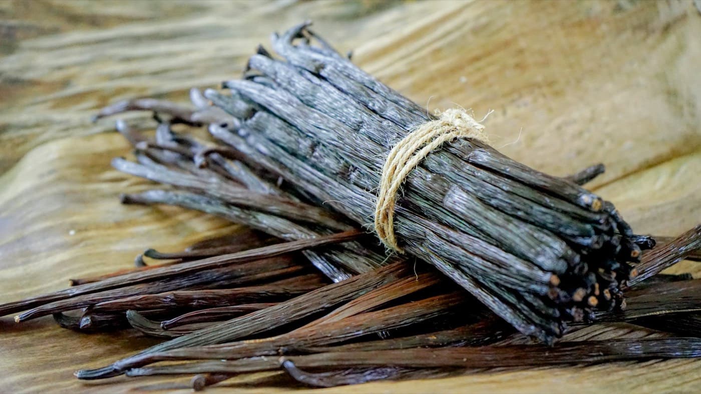 A bundle of cured vanilla beans tied with string, with loose beans beside it on a wooden board
