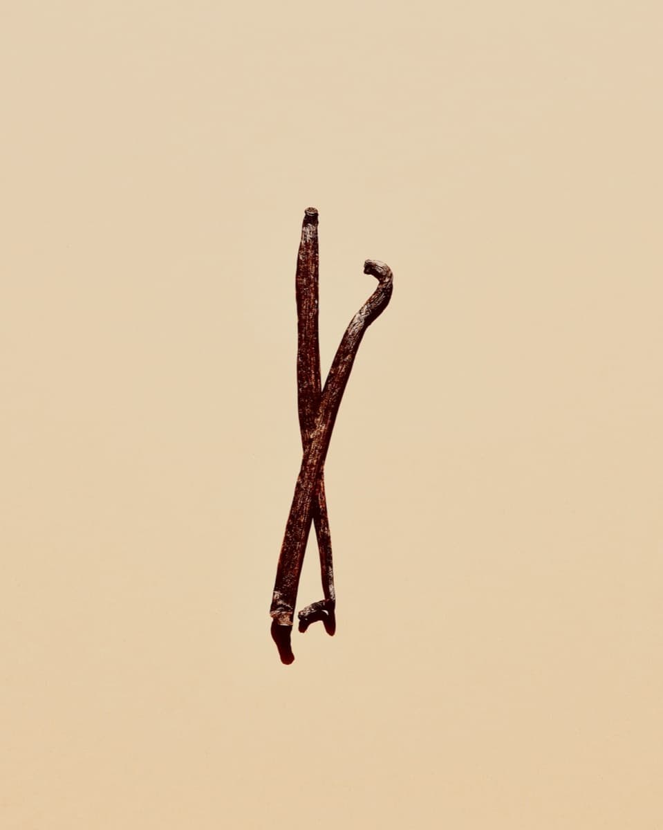 Two cured vanilla beans crossed on a pale cream background